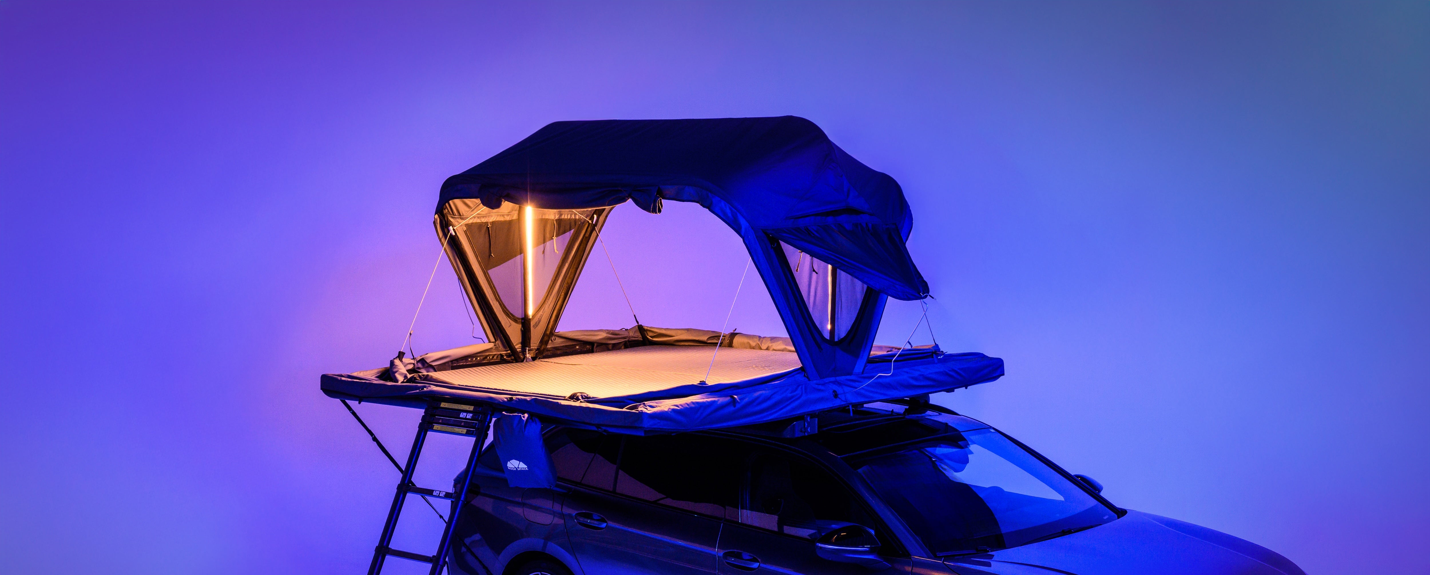Ein Auto mit einem beleuchteten Dachzelt in Blau- und Lilatönen. Eine Leiter führt zum Zelt. "Roof Space" ist auf einer Tasche sichtbar.