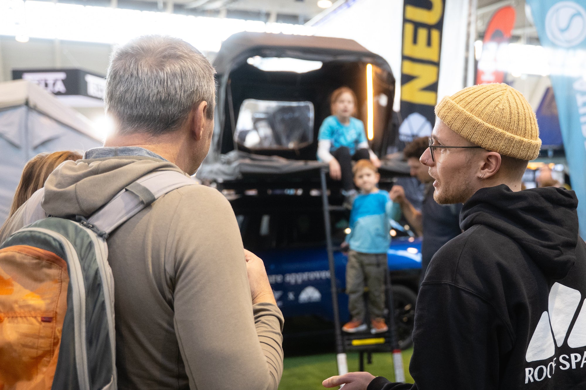 Aufnahme von einer Messe. Zwei Männer im Vordergrund unterhalten sich, im Hintergrund ein Fahrzeug mit Dachzelt und zwei Kinder. Auf dem schwarzen Hoodie steht "ROOF SPA".