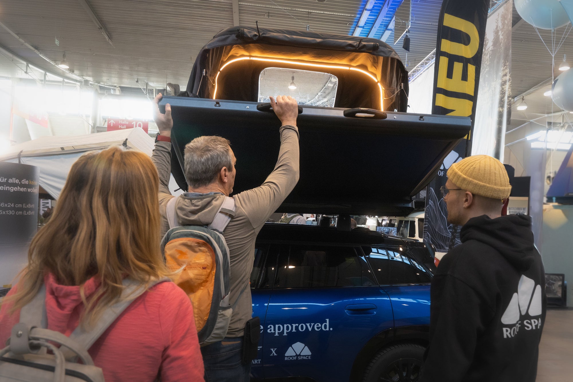 Auf einer Messe öffnet ein Mann ein Dachzelt auf einem blauen Auto; im Vordergrund stehen zwei weitere Personen. Im Hintergrund sieht man ein Banner mit der Aufschrift "NEU".