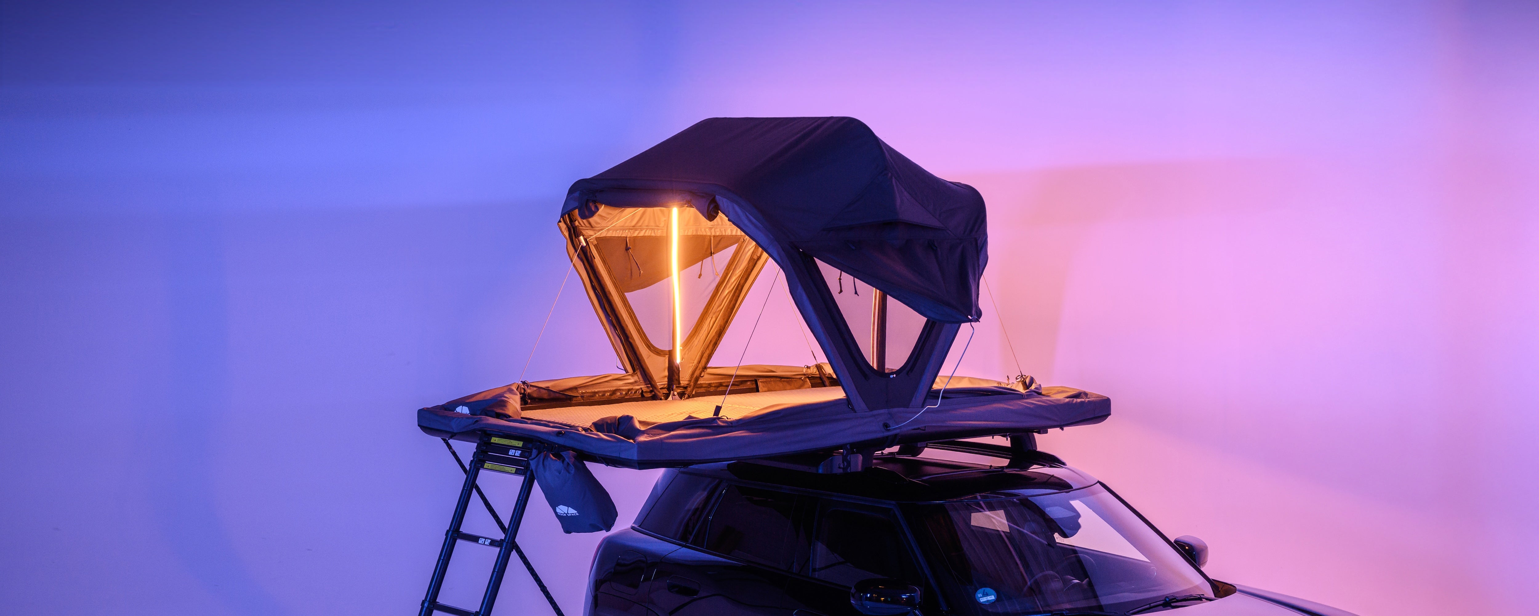 Ein Dachzelt in Schwarz auf einem Mini Cooper, beleuchtet mit warmem Licht, vor einem blau-violetten Hintergrund. Eine Leiter lehnt links an.